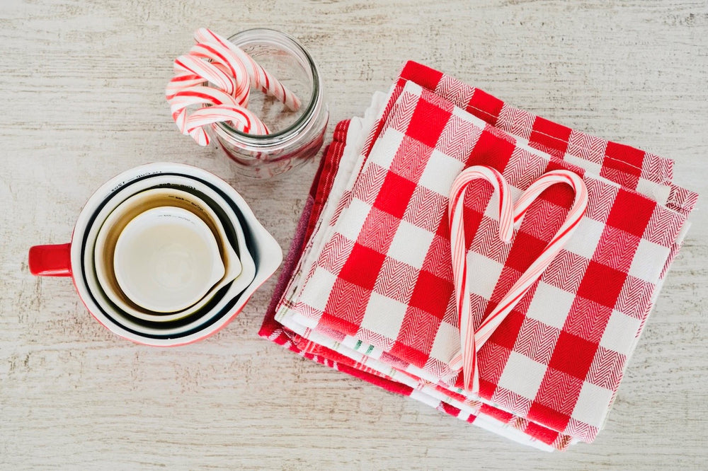 Candy Canes form the shape of a heart on red plaid napkin, with a stack of measuring cups and a glass of candy canes on a white wood table for Christmas In July